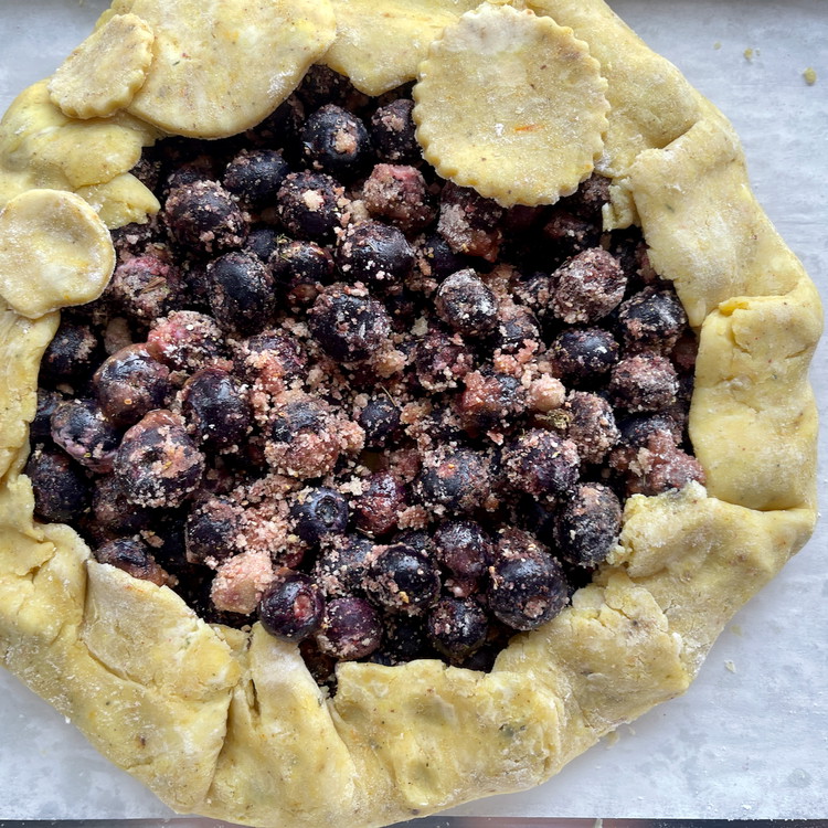 Image of Work around the edge of the galette, pushing the dough in to make a 1 ½” fold inward, tucking and overlapping as you continue around the edge. You can also lift the paper with the dough for extra support as...