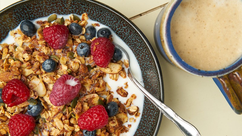 A bowl of granola and berries filled with milk, with foamed coffee to the side.
