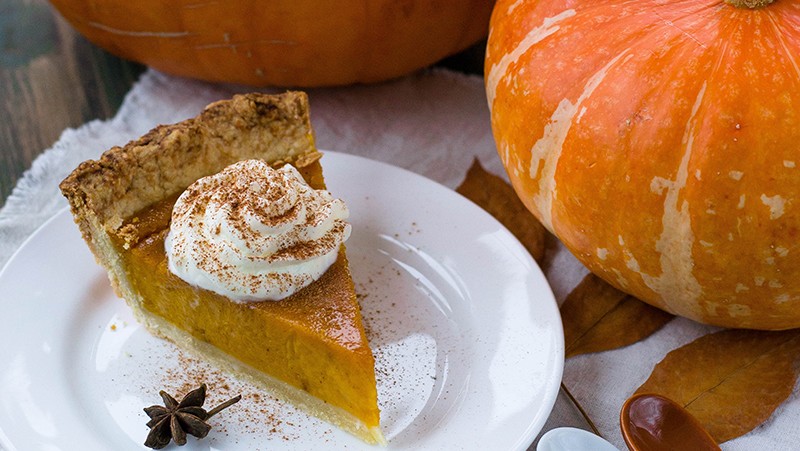 Piece of pumpkin pie on a plate with two large pumpkins on a table.
