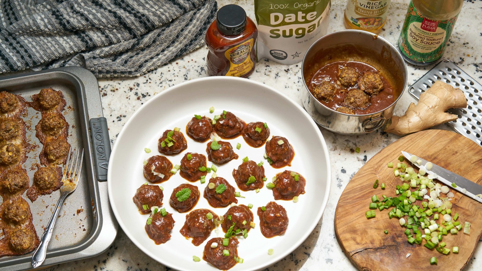 Tray of meatballs, meatballs in a bowl, meatball sauce, cut up chives, date sugar, and towels, all on a counter.