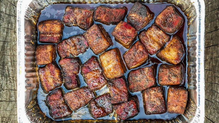 Image of Using tongs to transfer the pork belly cubes into a new aluminum pan. Add Blues Hog Original sauce to the pan and toss gently to coat. Return the pork belly cubes to the smoker uncovered for 5-10 minutes, just enough...