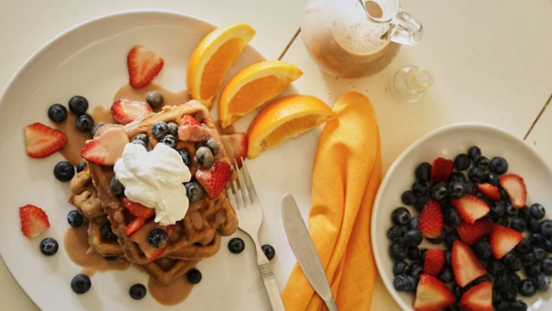 A person's hand drizzling syrup on a date sugar waffle plated with strawberries and blueberries. Purdate Sugar Recipe.