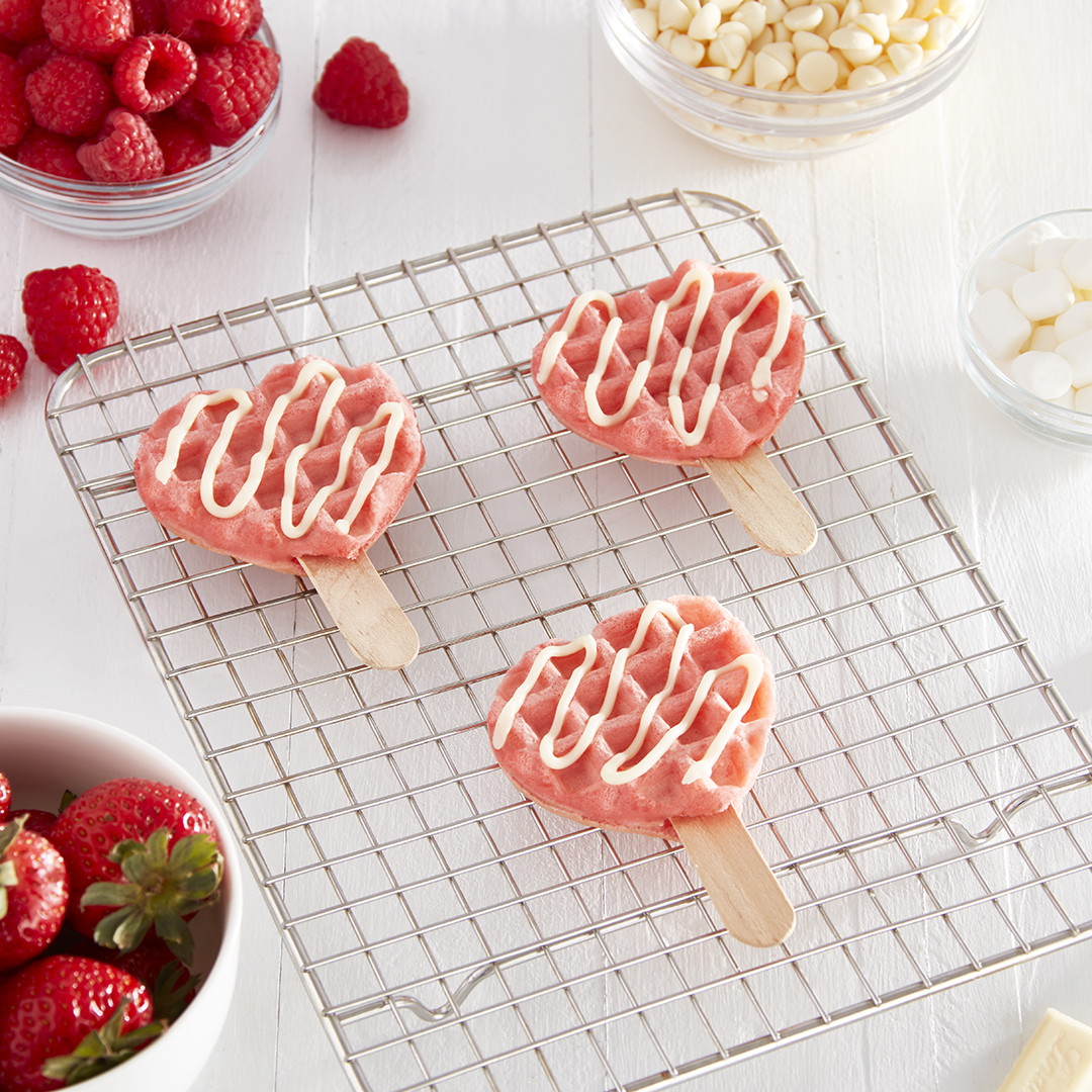 Three pink mini heart waffles with white frosting on ice pop sticks sit on a cooling rack.