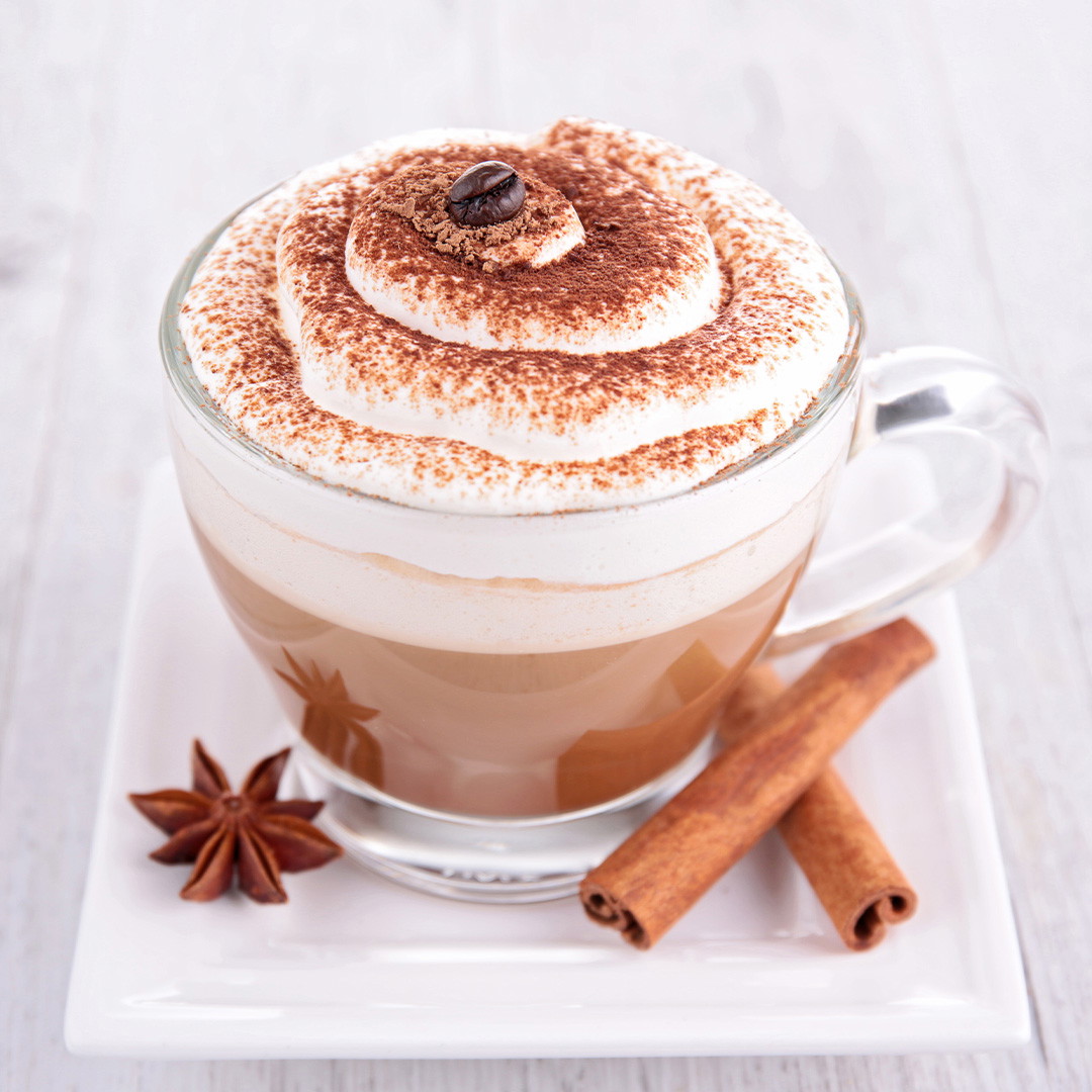 Café moka à la cannelle dans une tasse en verre, garni de crème fouettée et de cannelle, sur une assiette blanche.