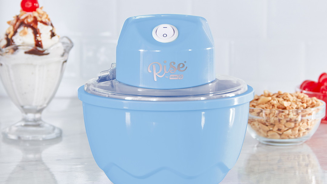 The Rise by Dash Personal Ice Cream Maker sits on a marble counter next to an ice cream sundae.