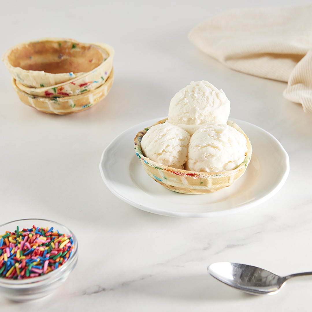 Un tazón de waffle con chispas de colores lleno con tres bolas de helado de vainilla sobre un plato blanco.