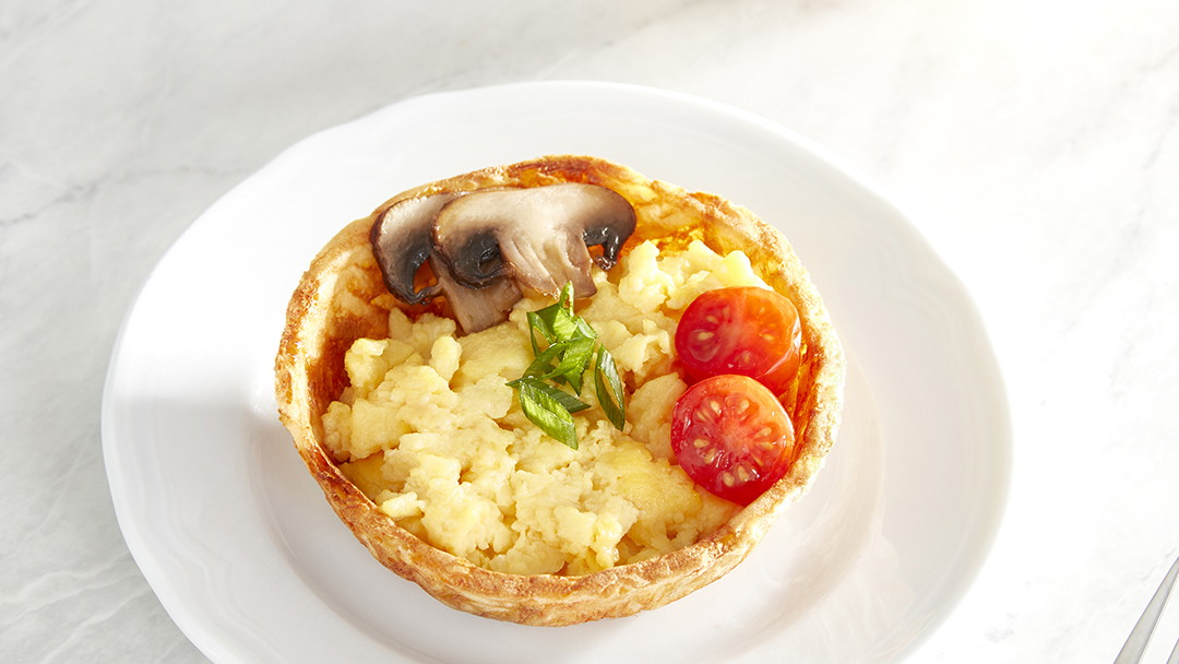 A hashbrown waffle bowl filled with scrambled eggs, tomato, and mushroom, garnished with chives, on a white plate.