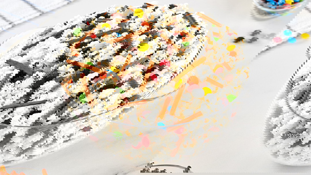 A clear bowl filled with popcorn, M&Ms candy, pretzel sticks and marshmellows.