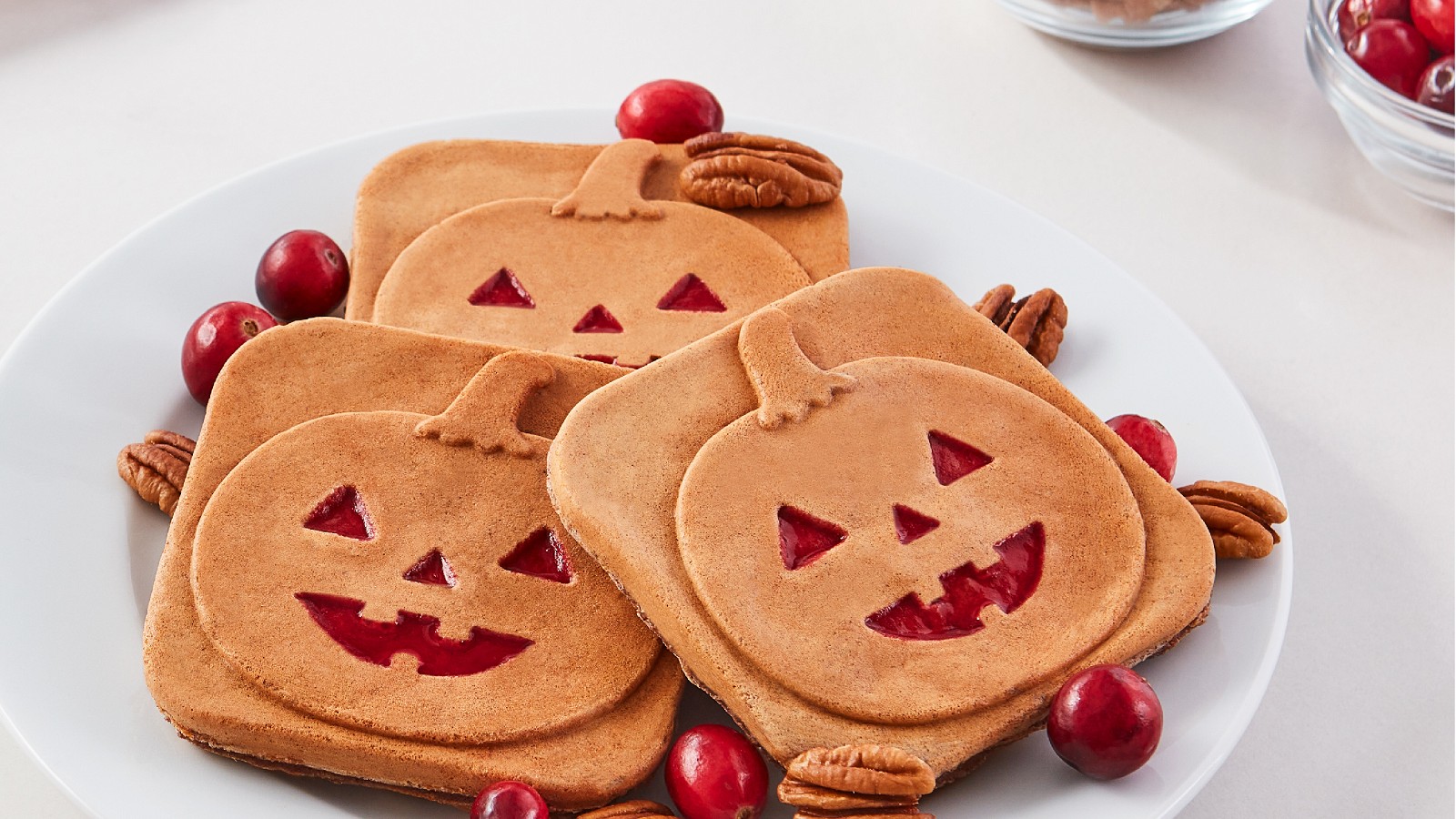 Three square mini waffles with a Jack-O-Lantern print on a white plate with pecans and cranberries.