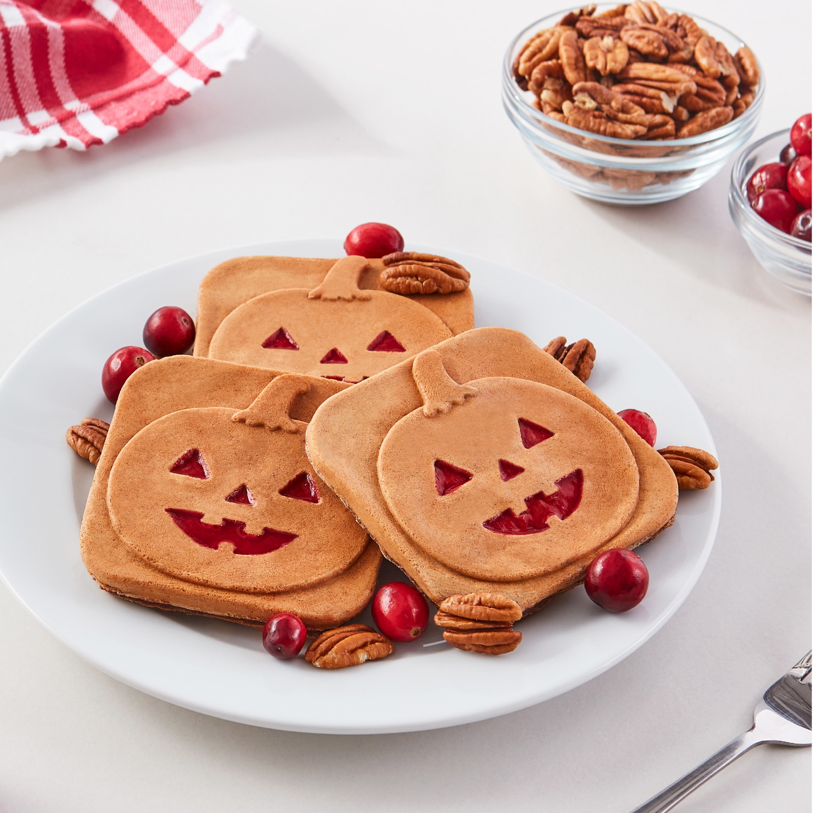 Trois mini gaufres carrées avec un motif de citrouille Jack-O'-Lantern sur une assiette blanche avec des pacanes et des canneberges.