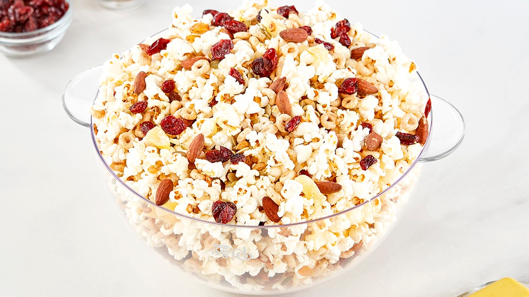 A clear bowl filled with popcorn, almonds, cranberries, and sunflower seeds.