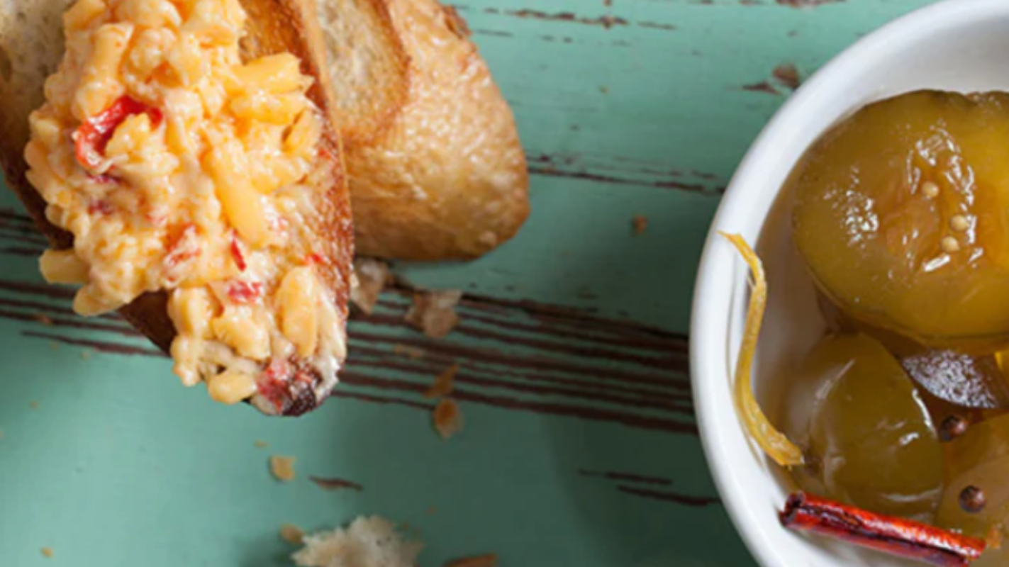 pickled pink pimento cheese on a toasted baguette slice, pickles in a bowl next to it