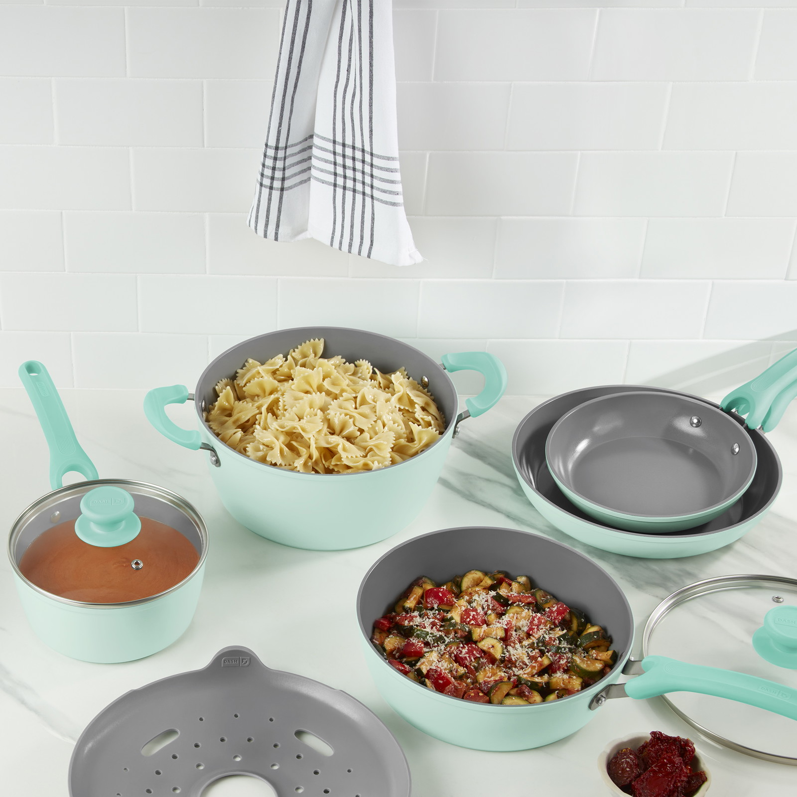 Un ensemble de casseroles et poêles sur un comptoir en marbre. Une grande casserole contient des pâtes papillon, la petite casserole contient de la sauce, et la poêle à frire profonde contient des tomates et des courgettes.
