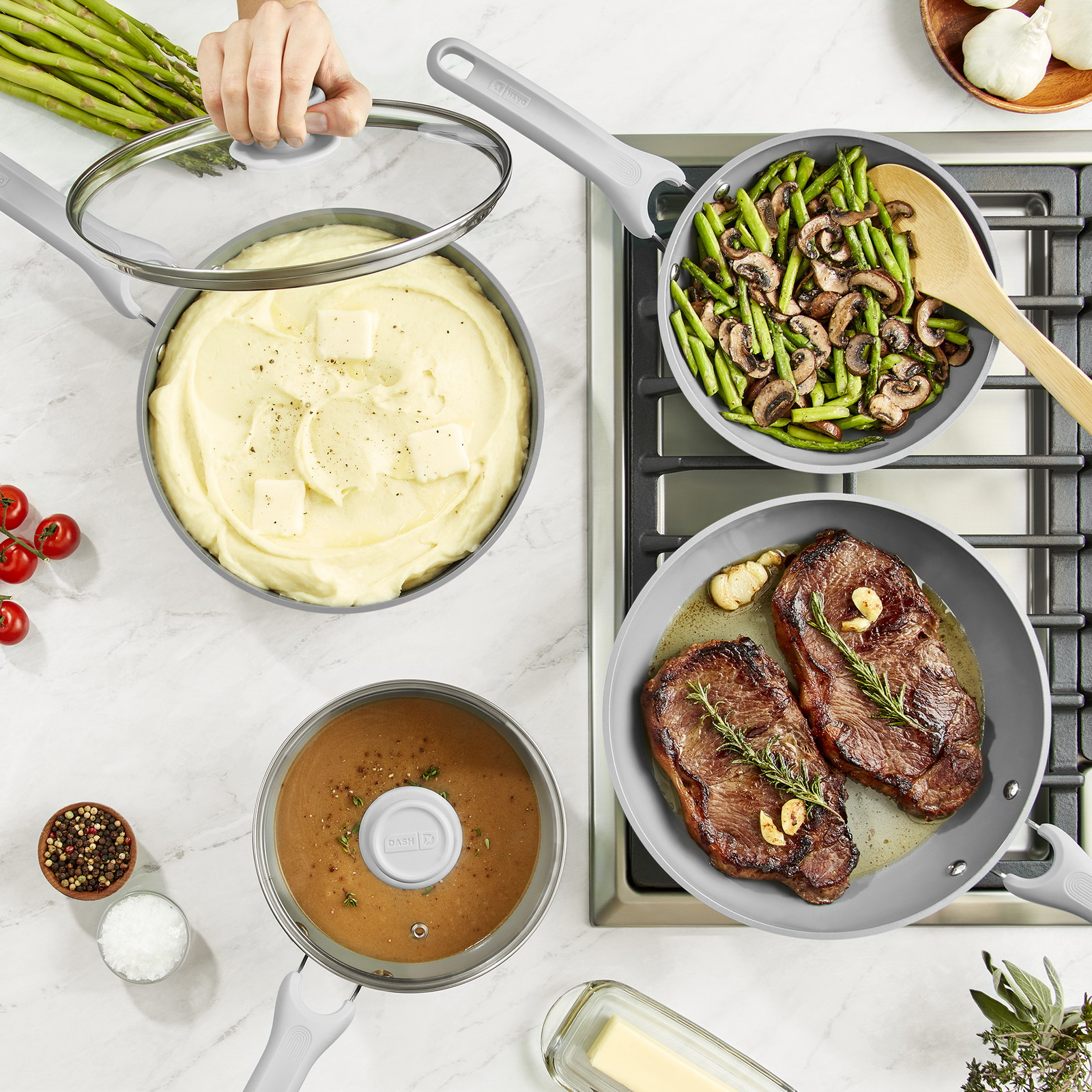 Vue de dessus de diverses casseroles et poêles. Deux poêles sur une cuisinière contiennent des steaks et des asperges aux champignons. Sur le comptoir se trouvent une casserole de purée de pommes de terre et une casserole de sauce.