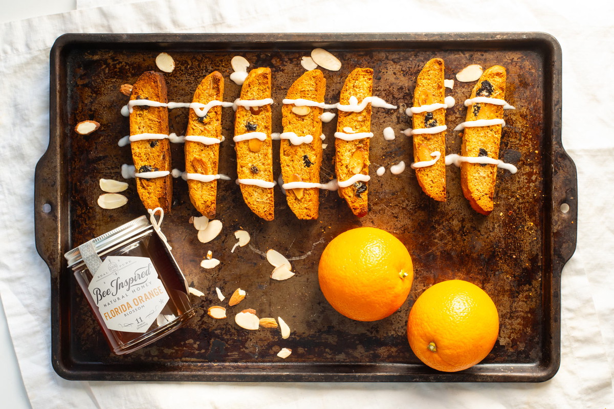 cranberry orange biscotti on top of a baking pan with bee inspired honey and whole oranges