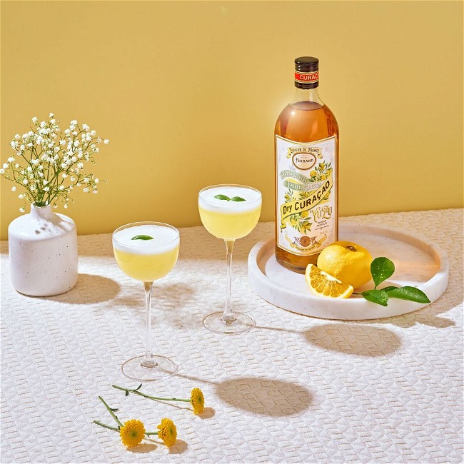 A tableau of beautiful objects: a bottle of Pierre Ferrand Dry Curacao Yuzu Late Harvest on a round serving tray, sliced lemons next to it. In the center, two cocktails with an elegant foamed top, a vase with a spray of baby's breath on the far left.