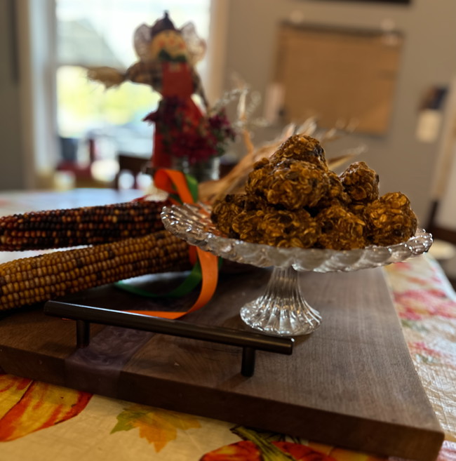 Sunny Pumpkin Balls placed in a pyramid on a candy plate placed onto of a butcher board. A bundle of color corn lays next to is with a mason jar of maroon mums and a small scarecrow sit behind them.