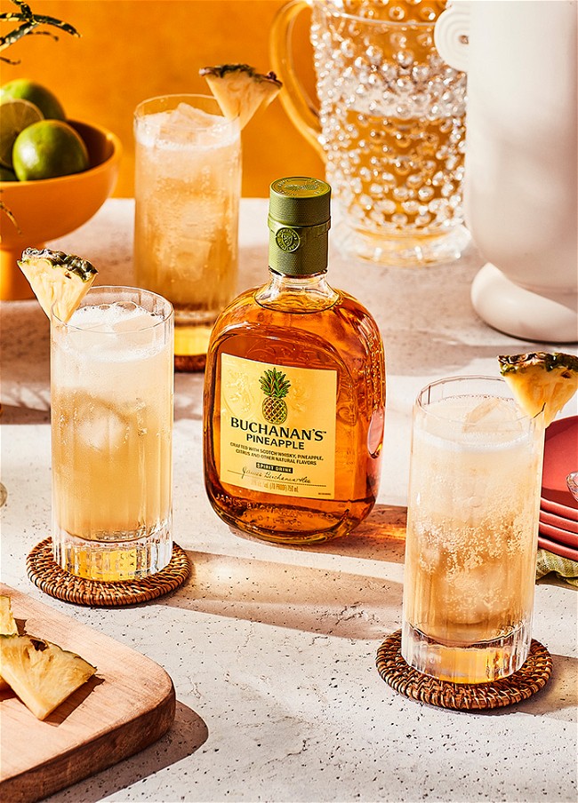 Two refreshing cocktails with pineapple wedge garnishes flanking a full bottle of Buchanan's Pineapple