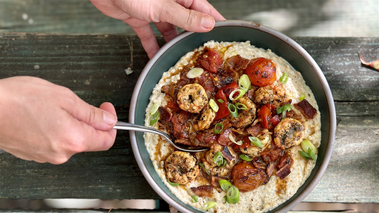 A bowl of creamy-looking grits topped with shrimp and sauce, with hands holding it and a wooden backdrop.