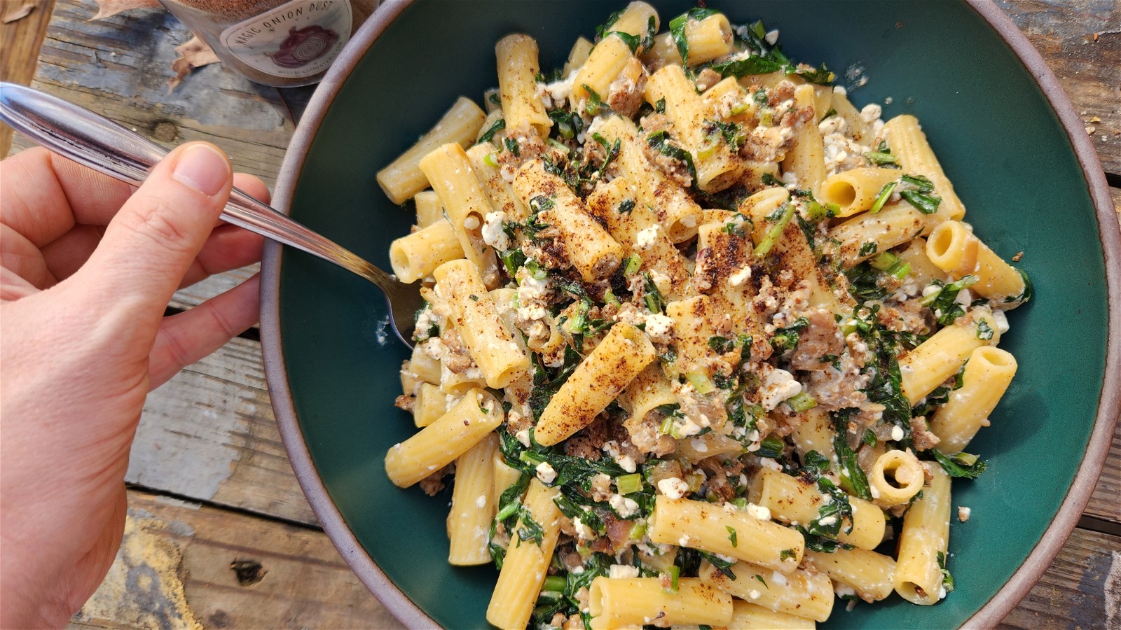 A teal ceramic bowl of creamy looking pasta with greens and sausage. A hand holds a spoon and it's on a wooden background.