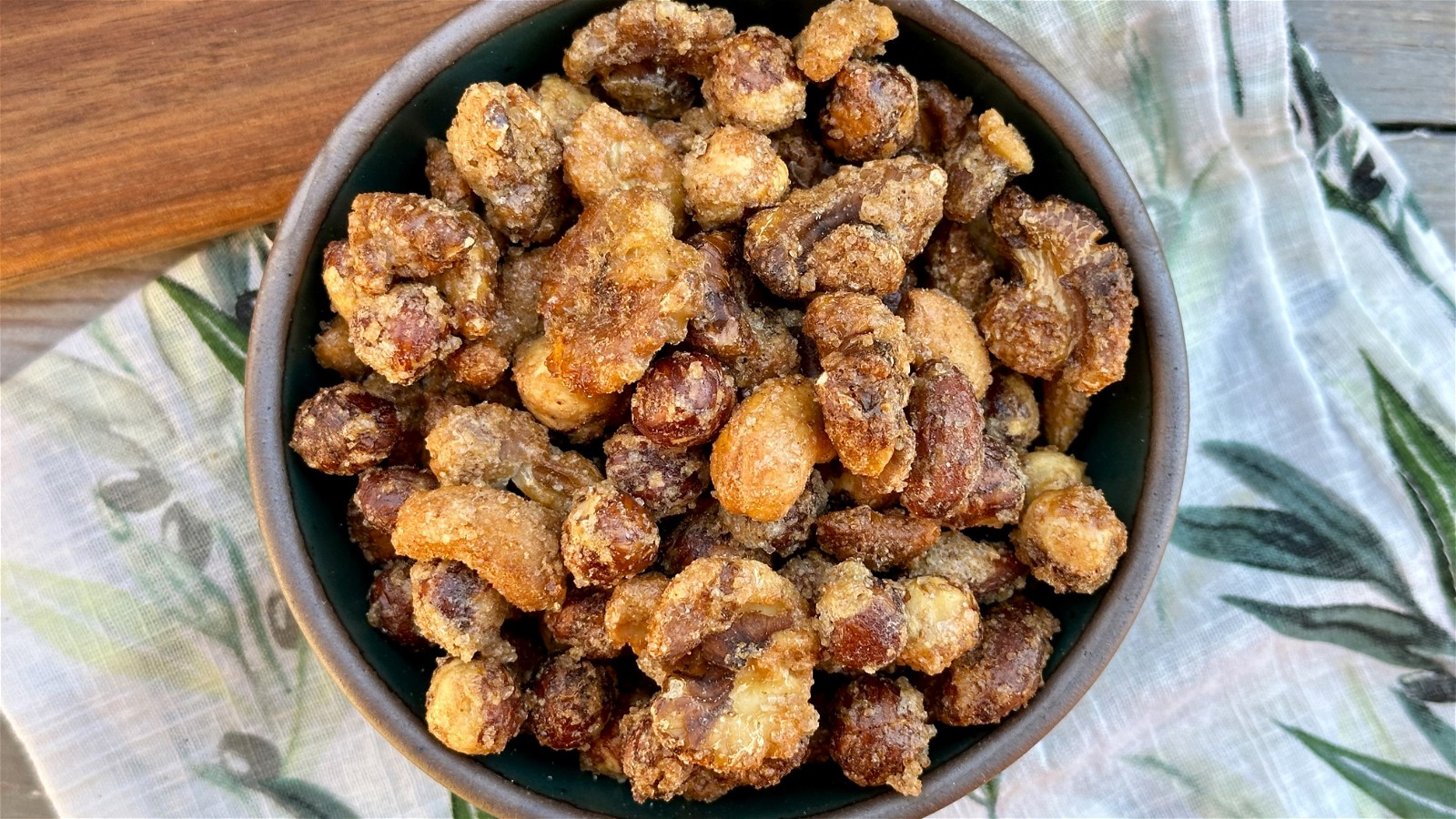 A ceramic bowl of candied mixed nuts on a wooden background with a fabric napkin.