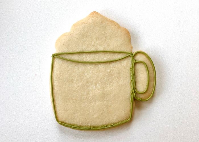 Image of Outline the shape of the mug with green piping consistency icing. Be sure to outline the rim of the glass and the handle of the mug as shown.
