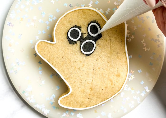 Image of Outline the entire cookie with white piping consistency icing. Pipe 3 circles around the dried black icing to create the eyes and mouth. Let the piping consistency icing dry for 10-15 minutes before moving on to the next step.