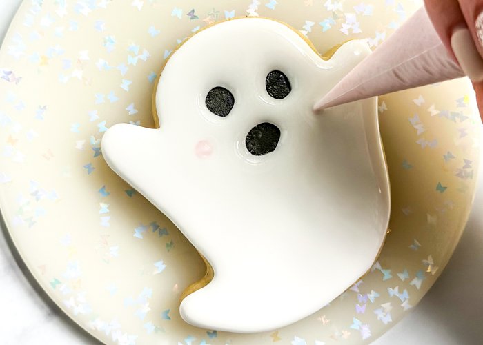 Image of While the white flooded base icing is still wet, pipe two small dots of light pink icing on the cheeks of the ghost. You could wait for the icing to dry to pipe the dots, this will make them stand out more. 