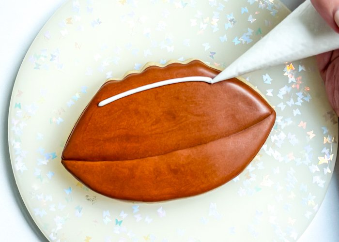 Image of Once the brown icing has crusted over, use white piping consistency icing to pipe a line on top of the football. This is where the laces will be.       Pro Tip:  When piping lines of piping consistency icing, it's important to position your hand slightly above the surface where you'll be starting your line, push out the icing and then let guide the icing where you want to start your line. Don't attempt to start your line with your tip touching the cookie. Apply even pressure and continue to guide the icing to lay where you'd like it to go. When you reach the end of your line, release the pressure and gently lay the end of the line down. This will create an even line without 