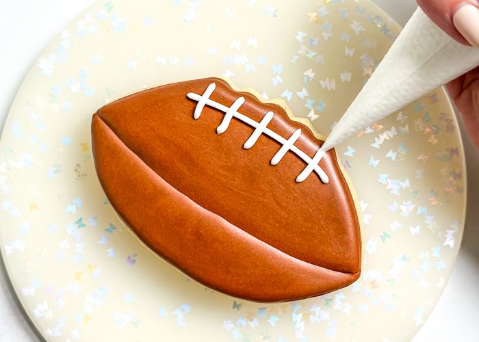 Image of Using white piping consistency icing, pipe 5-6 smaller lines perpendicular to the first line you piped, creating the football laces.