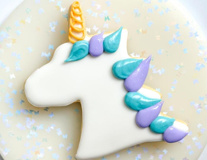 Image of Using the blue piping consistency royal icing, pipe the unicorn mane as shown in the photo. Use the tip of your bag to create a swooping look to the mane-it will create a mane that looks like it is flowing. Dispense royal icing with the tip of your piping bag pressed firmly on the cookie, then remove in a sweeping motion to get some movement in the mane.     Pro tip: It can be helpful to cut a larger hole than you would typically use for piping consistency so that you don't have to add more pressure to the bag and get a smoother effect to the icing. Let the icing dry for 30-60 minutes.