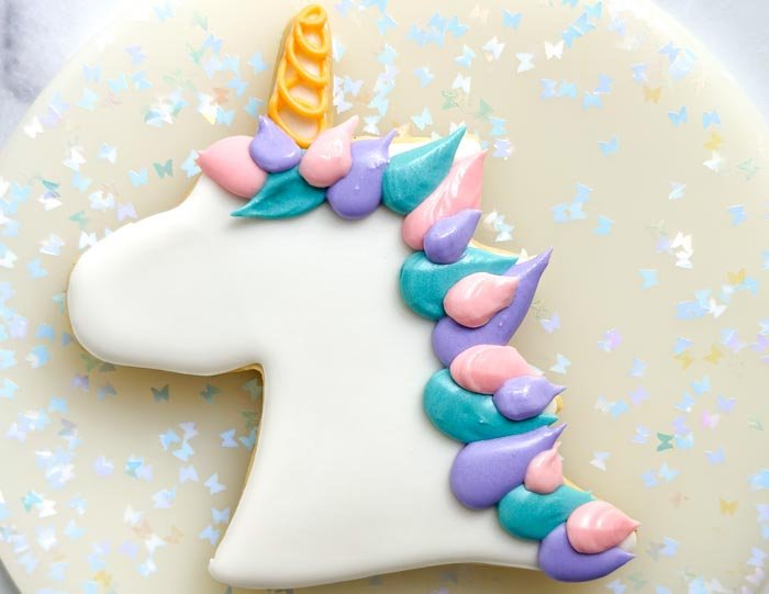 Image of Using the pink piping consistency royal icing, pipe the unicorn mane next to the purple icing you just piped. Let the icing dry for 30-60 minutes. At this point, you can stop if you feel like you have enough of a mane, or you can continue and do another layer of hair.