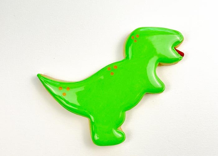 Image of While the flood icing is still wet, use piping consistency orange royal icing to pipe dots and swirls to give your dino some detail. Doing this step while your green flood icing is still wet will ensure that the details look like they are part of the dino, rather than standing out on the body of the dino. If you prefer more dimension, you can wait for your flood icing to dry 30-60 minutes before piping the details.
