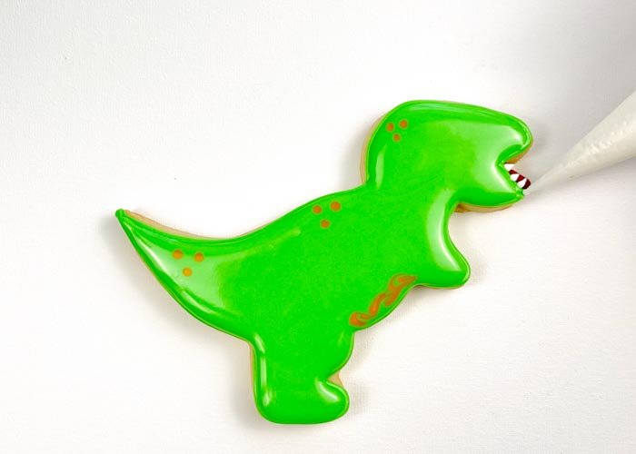 Image of Using white piping consistency royal icing, pipe dots on the top and bottom jaw to create dino teeth. To make them more pointy, gently drag the white icing with a toothpick or scribe tool.
