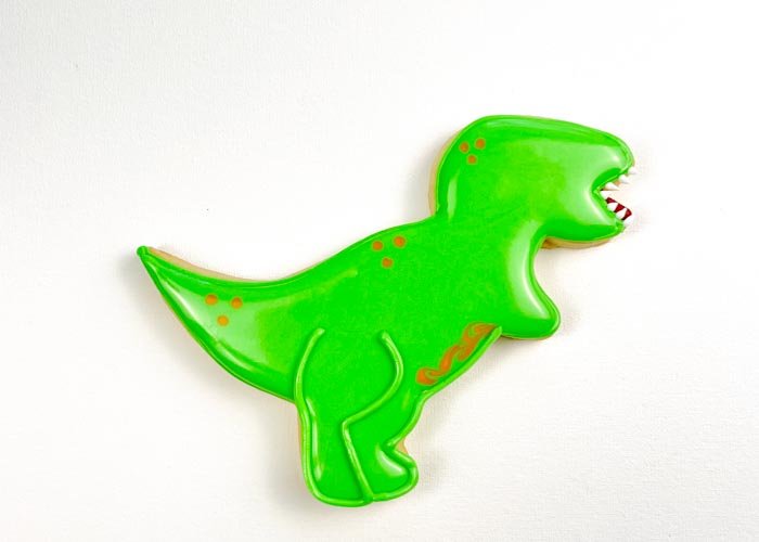 Image of Once your flood base has dried and begun crusting over, use green piping consistency icing to pipe fine lines on the dino to create differentiation between the arms and the legs. If you don't wait until the icing has dried, the piping consistency icing will blend in with the flood consistency icing, resulting in lines that aren't clearly defined.