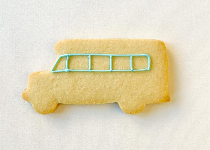 Image of Using piping consistency blue icing, outline the windows of the school bus, starting with the windshield. Be sure to pipe vertical lines to delineate between the individual windows.