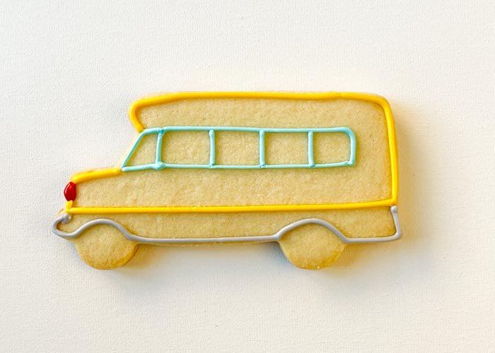 Image of Next, using yellow piping consistency icing, outline the main chassis of the school bus. While the yellow icing is still wet, adhere red candy for the headlight. Alternatively, you can use red royal icing to pipe the headlight. Alternatively, you can use a red candy or sprinkle to make the headlight. Use gray piping consistency icing to outline the lower part of the school bus, near the wheels.