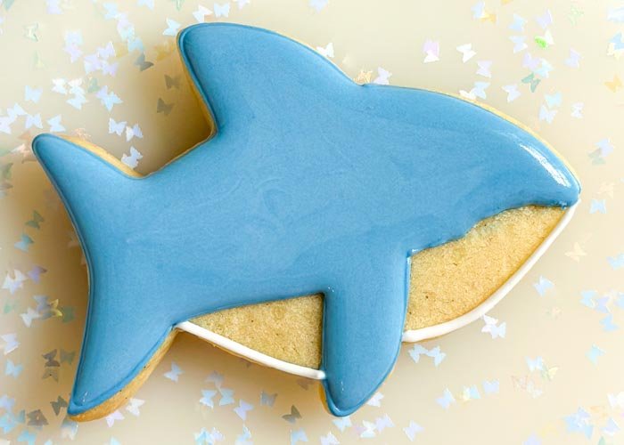 Image of Fill in the top portion of the baby shark with blue flood consistency royal icing. Let this part air dry for a minimum of 30 minutes before moving onto the next step. The icing should be crusted over-this will ensure that your colors won't bleed, and there will be good definition between the blue and white sections.