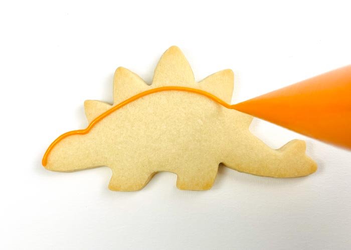 Image of Using piping consistency orange icing, outline the shape, omitting the spikes on the spine of the stegosaurus. Be sure to define all 4 legs when outlining, as this will help to add dimension to your stegosaurus.