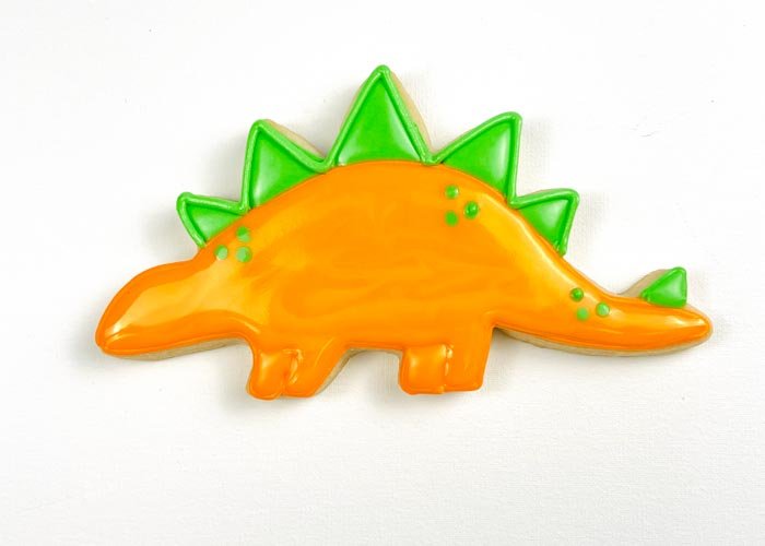 Image of While the flood icing is still wet, use piping consistency green royal icing to pipe dots to give your dino some detail. Doing this step while your orange flood icing is still wet will ensure that the details look like they are part of the dino, rather than standing out on the body of the dino. If you prefer more dimension, you can wait for your flood icing to dry 30-60 minutes before piping the details.