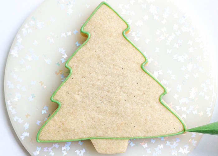 Image of Using the light green detail consistency icing, outline the outside of the cookie. Do not outline the trunk, only outline the main part of the tree.
