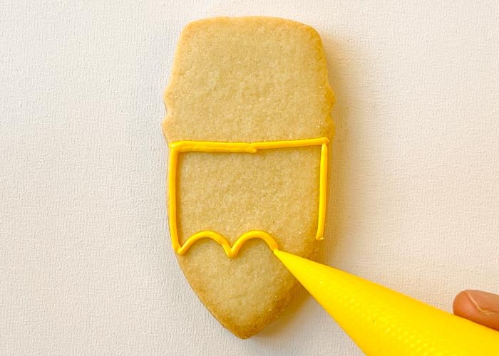 Image of Using piping consistency yellow icing, outline the pencil. Leave space at the top for the eraser and at the bottom for the pencil tip. Pipe a scalloped edge on the bottom to make your pencil look freshly sharpened.
