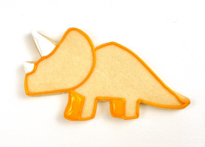 Image of Using piping consistency white icing, fill in the horns of the triceratops.  Using flood-consistency orange icing, fill the two small rectangles that will be the back legs of the triceratops. Wait for these to dry for about 10-15 minutes.