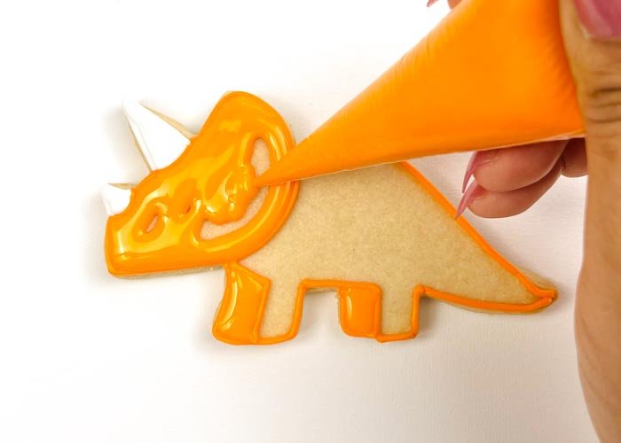 Image of Using piping consistency orange icing, pipe some squiggles in the head of the Triceratops. This will help to give your flood some structure, so that the icing doesn't collapse as it cools. Then, with orange flood consistency icing, fill in the head, using a scribe tool to make sure you have full coverage. Wait for the icing to dry for about 10-15 minutes.