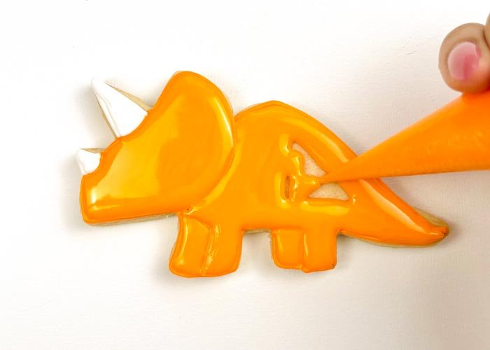 Image of Using piping consistency orange icing, pipe some squiggles in the body of the Triceratops. This will help to give your flood some structure, so that the icing doesn't collapse as it cools. Then, with orange flood consistency icing, fill in the body, using a scribe tool or toothpick to make sure you have full coverage.