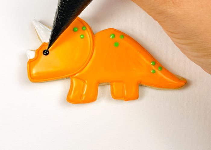 Image of Using black piping consistency royal icing, pipe a small dot to create an eye for your dino. If you want to skip mixing up a batch of black icing, you can also use a small candy or nonpareil to create the dinosaur eye.