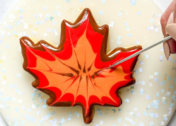 Image of While the brown, red, and orange icing is still wet, use a scribe tool or toothpick to gently pull the icing toward the edge of the leaf. Start from the middle of the leaf and drag the icing out. Repeat this process 10-15 times, until you're satisfied with the results. There is no need to clean your toothpick or scribe tool between each drag. The mixing of colors adds to the design, making a more realistic look. Let this base layer dry for 30-60 minutes, until the icing is just crusted over. You can place the cookies under or next to a fan to speed up the drying process.