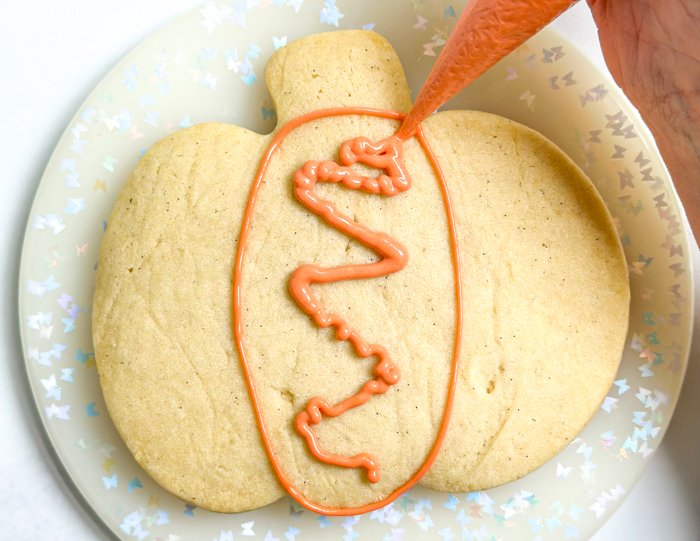 Image of Using the orange piping consistency icing, outline the middle of three sections of the pumpkin, as shown in the photo. Pipe a squiggle of the same icing in the middle of the section. Pro Tip: Piping a squiggle of outline consistency royal icing in the middle of small flood sections of a cookie help with creating a 