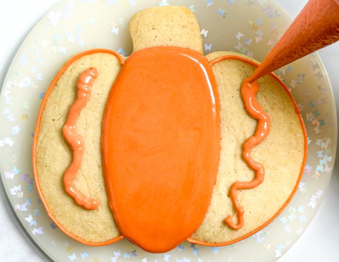 Image of Using the orange piping consistency icing, outline the outer two sections of the pumpkin. Pipe a squiggle of the same icing in the middle of the section. This will help to prevent the flood icing from collapsing and cratering as it dries.