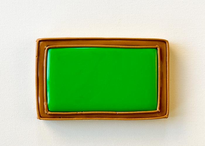 Image of Once flood icing has dried, use brown piping consistency icing to pipe a border around the edge of the cookie and around the green blackboard part of the cookie. This will give the frame of the blackboard some dimension.